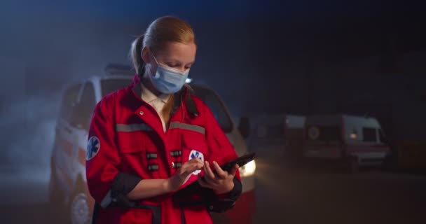 Kaukázusi fiatal női mentős piros egyenruhában és maszk légzőkészülék koppintás és gépelés a képernyőn tabletta számítógép. Női orvos kitölti az orvosi nyomtatványt az eszközön éjjel a szabadban. Vészhelyzet van. Kovid-19 — Stock videók