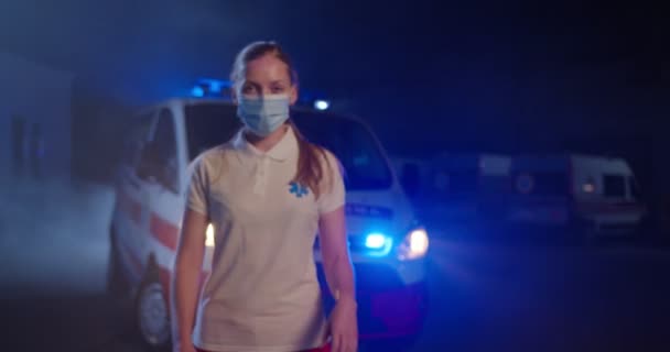 Retrato de paramédico femenino caucásico en máscara médica mirando a la cámara, cruzando las manos al aire libre. Ambulancia de fondo. Enfermera en turno de noche. Acercándote. Dolly disparó. pandemia de Covid-19. — Vídeos de Stock