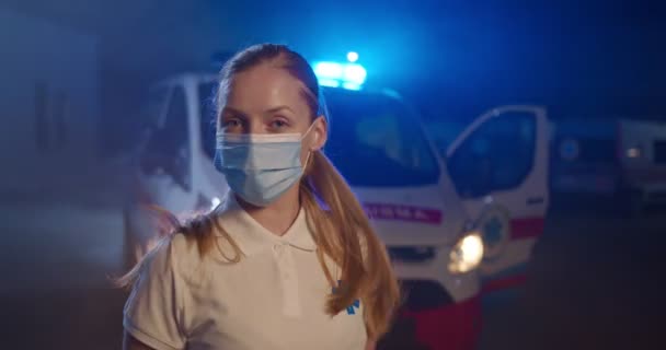 Ritratto di paramedica caucasica in maschera medica che guarda alla telecamera in strada. Ambulanza sullo sfondo. Infermiera al turno di notte. Chiamata d'emergenza. Pandemia di coronavirus. Aiuto paramedico. — Video Stock