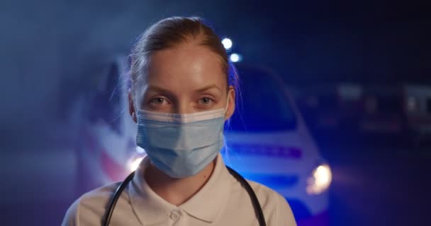 Gros plan du visage ambulancier féminin caucasien dans un masque médical regardant la caméra dans la rue. Ambulance en arrière-plan. Infirmière de nuit. Urgence. Pandémie de coronavirus. Aide paramédicale Portrait — Video