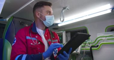 Beyaz, yakışıklı, kırmızı üniformalı, eldivenli, tıbbi maskeli, ambulansta oturan ve tablet kullanan bir sağlık görevlisi. Erkek doktor gece vardiyasında çalışıyor ve bilgisayarı tıkırdatıyor ya da kaydırıyor.
