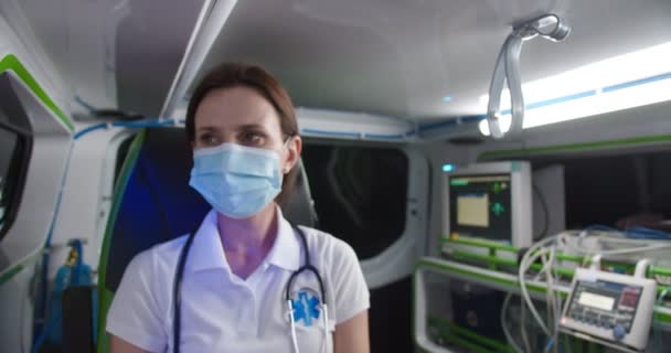 Primer plano de la cara paramédica femenina caucásica en máscara médica mirando a la cámara dentro de la ambulancia. Interior del coche. Enfermera en turno de noche. Emergencia. pandemia de Coronavirus. Ayuda paramédica. Retrato. — Vídeo de stock