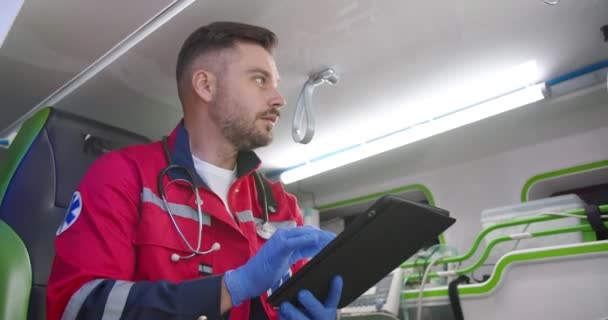 Paramédico masculino guapo joven caucásico en uniforme rojo y guantes sentados en ambulancia y usando dispositivo de tableta. Médico serio trabajando en turno de noche y tocando o desplazándose en el ordenador. — Vídeos de Stock