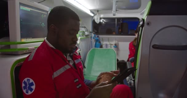 Afro-americano paramédico do sexo masculino sentado em ambulância e usando dispositivo tablet. Homem médico trabalhando no turno da noite e tocando ou rolando no computador. Médica caucasiana no fundo. Colegas. — Vídeo de Stock