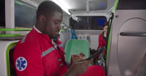 Afro-americano paramédico do sexo masculino sentado em ambulância e usando dispositivo tablet. Médico homem trabalhando no turno da noite e tocando ou conversando no computador. Médica caucasiana no fundo. Paramédicos. — Vídeo de Stock