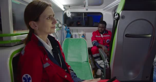 Paramédico femenino caucásico sentado en ambulancia e yendo a la paciente. Médico mujer trabajando en el turno de noche. Médico afroamericano de fondo. Paramédicos colegas. — Vídeo de stock