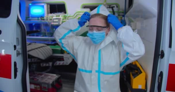 疲惫、疲惫的女医生，戴着个人防护设备和医疗面罩，摘下头罩和眼镜，坐在救护车上休息。PPE中女性医生的画像。Coronavirus概念. — 图库视频影像