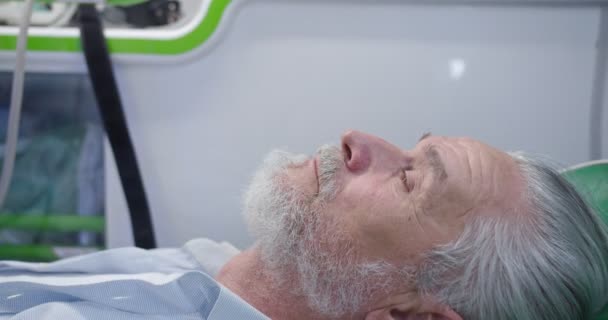 Крупный план белого старика с седой бородой, лежащего в машине скорой помощи с закрытыми глазами. Коронавирус. Врач дает кислородную маску пациенту. Реанимация. Работа в больнице. Проблемы с дыханием. Ковид-19. — стоковое видео