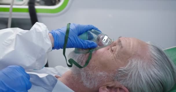 Primo piano del vecchio caucasico in ambulanza con gli occhi chiusi. Coronavirus. Il dottore dà la maschera di ossigeno al paziente maschio. Rianimazione. Ospedale che rianima il lavoro. Problemi respiratori. Salvare la vita durante Covid — Video Stock