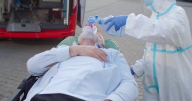 Kişisel koruyucu ekipman sağlıkçısı oksijen maskeli ihtiyarı ambulansa götürüyor. Coronavirus salgını konsepti. PPE 'de sağlık görevlisi. Doktor ve erkek hasta. Kapsamlı semptomlar. Dışarıda..