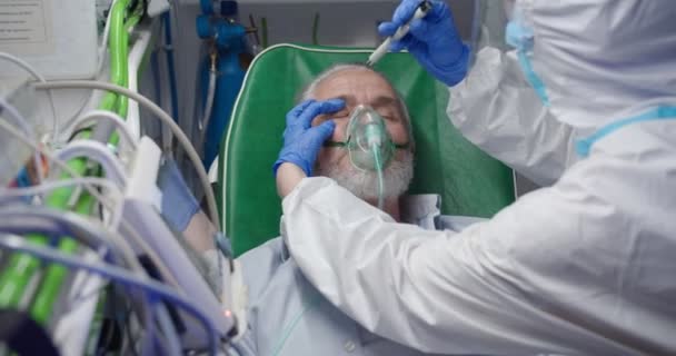 Arzt in persönlicher Schutzausrüstung hilft einem alten Mann im Krankenwagen. Sanitäter geben dem Patienten Sauerstoff und kontrollieren die Reaktion der Augen. Reanimation. Medizinische Arbeit während der Schwangerschaft. Erste Hilfe von Sanitätern. PSA. — Stockvideo