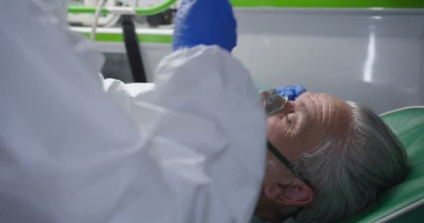 Detailní záběr zdravotníka v osobních ochranných pomůckách, jak zachraňuje staříka v sanitce. Medik dává kyslík pacientovi a kontroluje reakci očí. Oživení. První pomoc od doktora. PPE. Pacient s kovidem. — Stock video