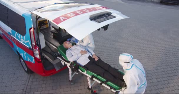 Two medics in personal protective equipment bringing old man in oxygen mask to the ambulance. Coronavirus pandemic. Paramedics in PPE. Doctors with male senior patient. Covid-19. — Stock Video