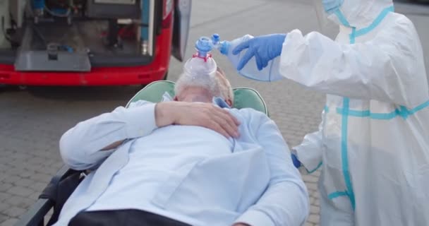 Sanitäter in persönlicher Schutzausrüstung bringen einen alten Mann mit Sauerstoffmaske zum Krankenwagen. Coronavirus-Pandemiekonzept. Sanitäter in der PSA. Arzt mit Patient. Covid sympthoms. Draußen. — Stockvideo