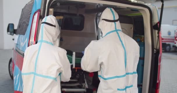 Male and female happy medics in personal protective equipment putting empty bed in ambulance car and going away cheerfully. Outside. End of working day. Covid-19 concept. Doctors in protection. — Stock Video