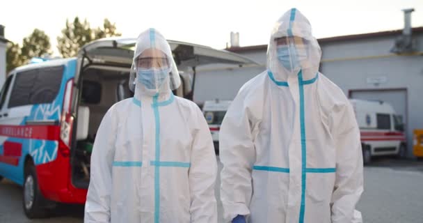 Portrait of male and female medics in personal protective equipment standing outdoors at ambulance and looking at camera. Colleagues paramedics in PPE. Doctors in protection during coronavirus. — Stock Video