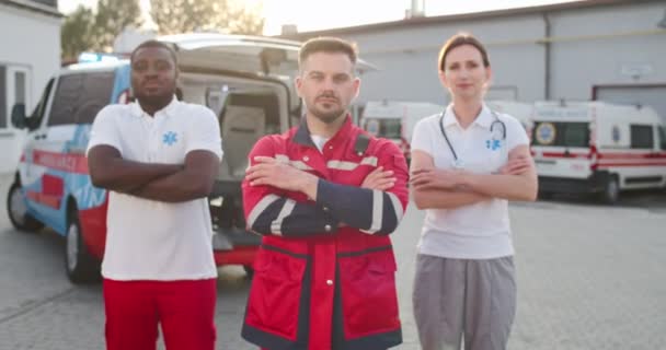 Portrait of mixed-races young team of paramedics in uniform standing outdoor with crossed hands and looking at camera. Ambulance on background. Multiethnic males and female doctors. Medics at work. — Stock Video