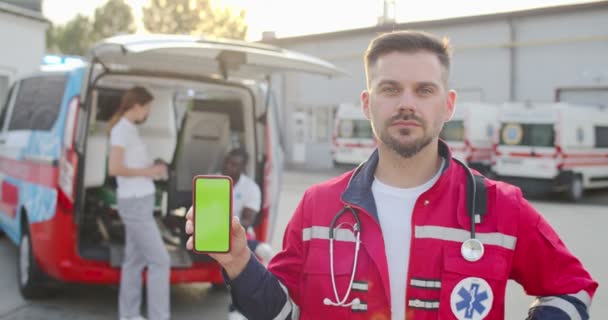 Ritratto di giovane uomo caucasico medico che guarda la fotocamera e mostra smartphone con schermo verde all'aperto. Ambulanza con paramedici in background. Medico di sesso maschile con cellulare in mano. Chiave cromata. — Video Stock