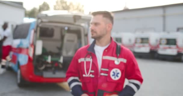 Porträtt av ung vit man sjukvårdare i röd uniform vänder leende ansikte och tittar på kameran utomhus. Ambulans på bakgrunden. Manlig stilig läkare. Oklar. Zoomar in. Dolly skjuten. — Stockvideo