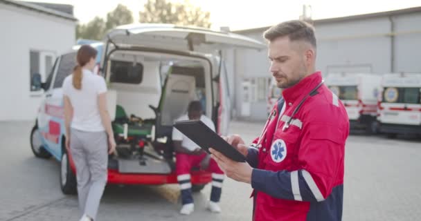 Portrait de jeune homme caucasien médecin regardant la caméra et utilisant un appareil tablette à l'extérieur. Des collègues ambulanciers en arrière-plan. Homme médecin tenant l'ordinateur et tapotant dessus. — Video