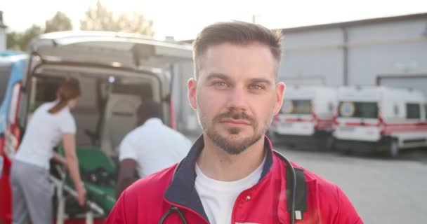 Retrato de un joven médico caucásico en uniforme rojo mirando a la cámara con la cara feliz sonriente al aire libre. Ambulancia con colegas de trabajo paramédicos de fondo. Hombre guapo doctor. De cerca.. — Vídeos de Stock