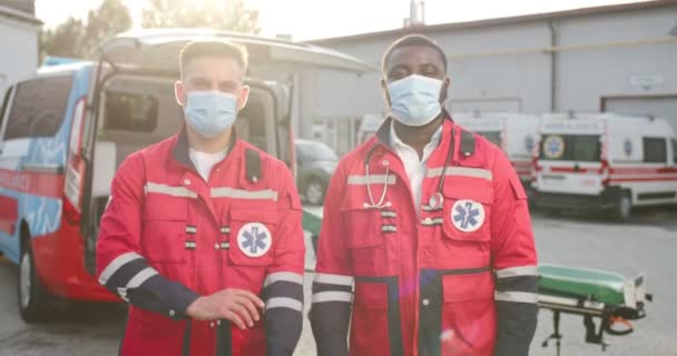 Portrét dvou smíšených mladých mužů v uniformách a maskách stojících venku se zkříženýma rukama a hledících do kamery. Záchranka na pozadí. Multietničtí doktoři. Zdravotníci v práci. — Stock video