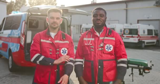 Porträtt av två blandade raser unga män paramedicin i uniformer står utanför med korsade händer och tittar på kameran. Ambulans på bakgrunden. Multietniska män läkare. Läkarvänner på jobbet. — Stockvideo