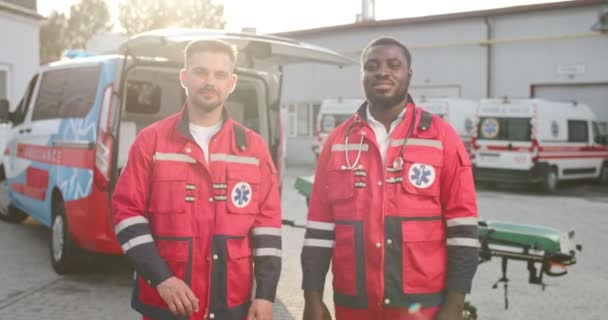 Portrait of two mixed-races young friendly males paramedics in uniforms standing outdoors and looking at camera. Ambulance on background. Multiethnic men doctors. Medics friends at work. — Stock Video