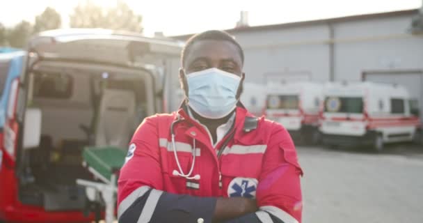 Portrét mladého afroamerického zdravotníka v červené uniformě a s maskou hledícího na kameru venku. Záchranka na pozadí. Hezký mužský doktor. Přibližuji. Dolly postřelená. Koronavirus. — Stock video