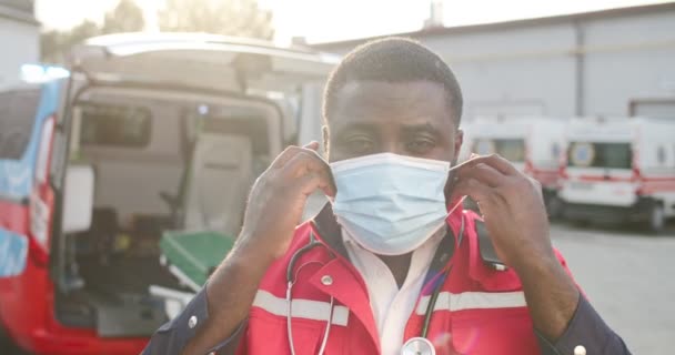 Ritratto di giovane afroamericano in uniforme rossa che indossa una maschera e guarda la telecamera all'aperto. Ambulanza sullo sfondo. Un bel dottore maschio. Protezione Covid-19. — Video Stock