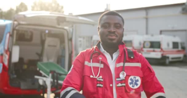 Retrato de jovem afro-americano médico em uniforme vermelho virando rosto sorridente e olhando para a câmera ao ar livre. Ambulância no fundo. Médico bonito. Desfocado. A aproximar-se. Dolly atirou. — Vídeo de Stock