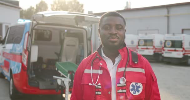 Porträtt av ung afroamerikansk man sjukvårdare i röd uniform ler och tittar på kameran utomhus. Ambulans på bakgrunden. Manlig stilig läkare. Sjukvårdare vid dagskiftet. — Stockvideo