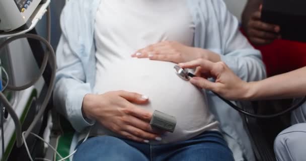 Close-up van blanke vroedvrouw met stethoscoop luisterende hartslag van baby in de buik. Zwangere vrouw in weeën op weg naar het ziekenhuis in een ambulance. Weeën. Bevallen van een kind. — Stockvideo