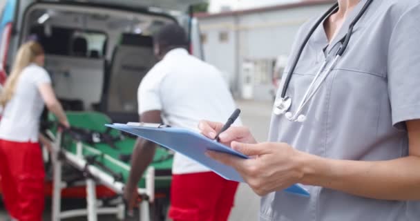 Primer plano de las manos femeninas del médico que rellenan el formulario médico y registran a un nuevo paciente de ambulancia. Al aire libre. Médicos de razas mixtas o enfermeras que trabajan en el coche en segundo plano. Escribir en documento. — Vídeos de Stock