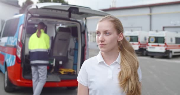 Portret van een jonge blanke vrouw die glimlachte en naar de camera keek. Ambulance op de achtergrond met artsen aan het werk. Vrouwelijke mooie dokter. Blond verpleegster. — Stockvideo
