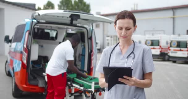 Kvinnlig läkare fyller i medicinsk form och använder bordsutrustning. Utomhus. African American doc arbetar på ambulans på bakgrund. Kvinnlig läkare knacka och bläddra på datorn på nätet. Läkarteam. — Stockvideo