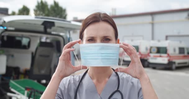Πορτάτο νεαρής Καυκάσιας όμορφης γυναίκας γιατρού που στέκεται έξω στο ασθενοφόρο και φοράει ιατρική μάσκα. Όμορφη γυναίκα γιατρός φορώντας αναπνευστήρα και κοιτάζοντας την κάμερα. — Αρχείο Βίντεο