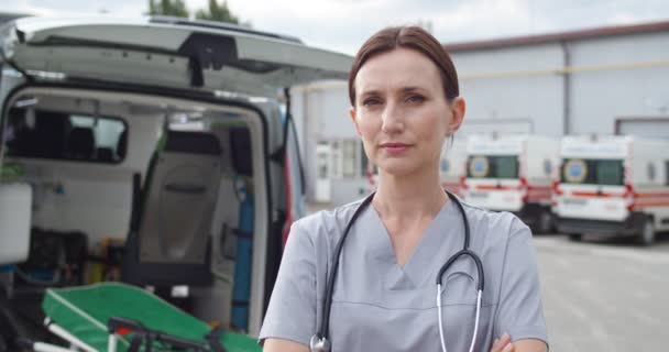 Ritratto di giovane donna caucasica bella medico guardando la macchina fotografica all'aperto. Aprire l'ambulanza sullo sfondo. Bella dottoressa con stetoscopio. Da vicino.. — Video Stock
