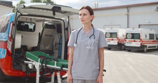 Porträtt bild av vackra unga kvinna läkare står utanför framför kameran och vänder ansiktet. Första hjälpen-konceptet. Ambulans på bakgrunden. Kvinnlig läkare i uniform och med stetoskop. — Stockvideo