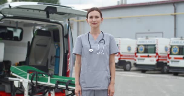 Portret van een mooie jonge vrouwelijke arts die voor de camera staat en glimlacht. Eerste concept van medische hulp. Ambulances op de achtergrond. Vrouwelijke arts met stethoscoop. Ziekenhuis. — Stockvideo