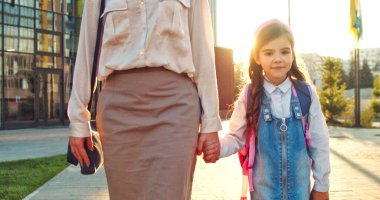 Beyaz bir kadın küçük bir kızın elini tutuyor ve sokakta okula yürüyerek gidiyor. Yeni yetme kız öğrencinin portresi annemle güneş ışığında derslere gidiyor. Okula dönelim. Konsept çalış