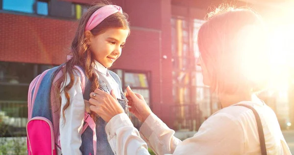 Close up retrato de caucasiano bela mulher fixando roupas em bonito pequena filha ao ar livre em luzes do sol. Vista lateral da mãe feliz enviar aluno menina para a escola na manhã ensolarada De volta ao conceito de escola — Fotografia de Stock