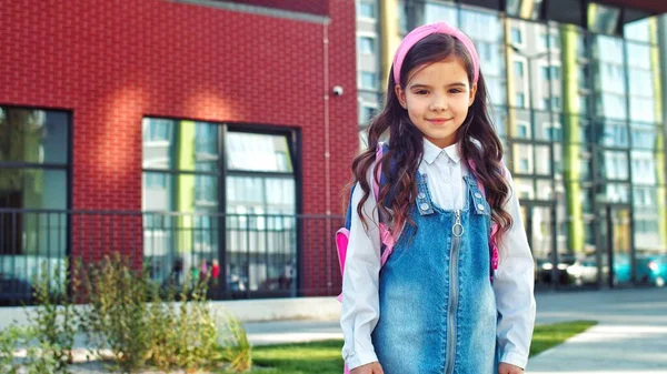 Retrato de feliz bonito linda menina da escola em rosa hairband sorrindo enquanto está de pé no quintal da escola. Criança bonita de bom humor ao ar livre. Estudante júnior. Conceito de educação — Fotografia de Stock