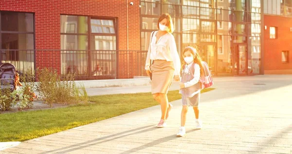 Mãe feliz caucasiana e pequena colegial bonito em máscaras andando ao ar livre no dia ensolarado da escola e pulando. Uma mulher muito alegre com um miúdo a correr e a sorrir na cidade. Conceito de quarentena — Fotografia de Stock