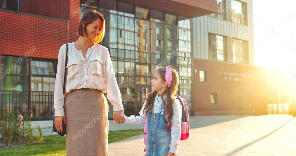 Caucasian cheerful woman holding hand of little daughter and standing near school outdoors on street. Happy schoolgirl holds mother hand in sun lights and smiling to camera. Back to school concept