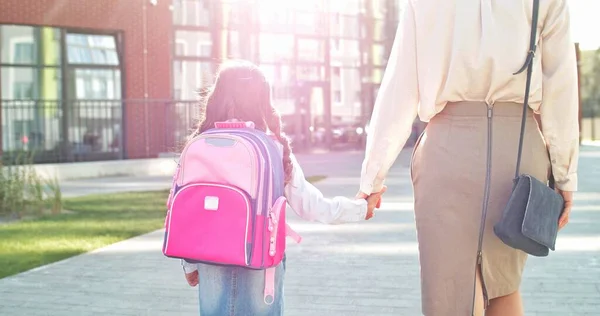 Traseira de caucasiano alegre menina bonito com mochila rosa indo com a mãe para a escola. Linda pequena estudante júnior do sexo feminino correndo para a escola de bom humor. De volta à escola. Conceito familiar — Fotografia de Stock