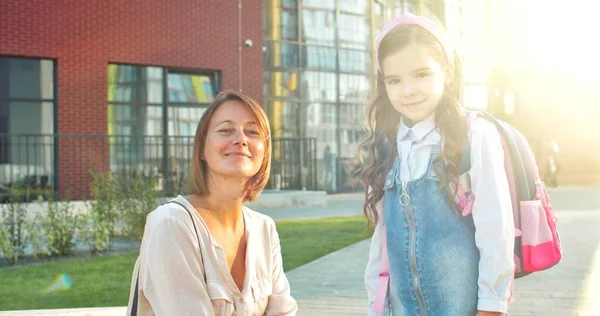 Retrato de mãe e filha caucasiana feliz sorrindo para a câmera perto da escola ao ar livre no dia ensolarado. Mulher alegre bonita com colegial bonitinho no pátio da escola após as aulas. Conceito de educação — Fotografia de Stock