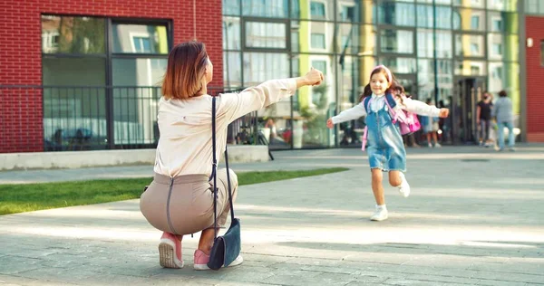 Вид сзади: красивая женщина встречает симпатичную школьницу из школы после уроков. Крупным планом радостный ребенок ученик с розовым рюкзаком бежит к матери и обнимает ее. Семейная концепция — стоковое фото