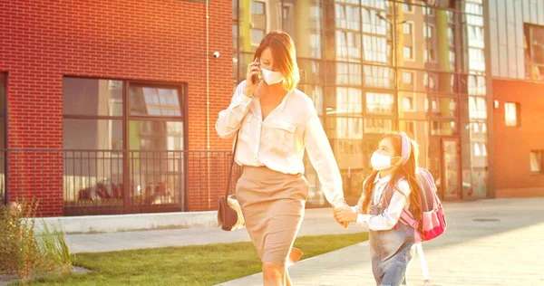 Mulher ocupada caucasiana em máscara falando no smartphone enquanto caminhava com a filha bonita da escola no dia ensolarado. Menina tentando chamar a atenção das mães que está chamando o conceito de escola de celular — Fotografia de Stock