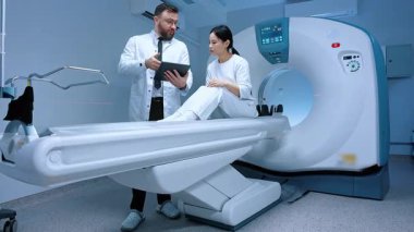 MR çekilmeden önce doktor hastayla konuşuyor. Doktor tableti tutuyor. Hasta tomografide oturuyor. Doktor MRI işleminden önce talimat veriyor. Manyetik rezonans görüntüleme. Doktor ve hasta..